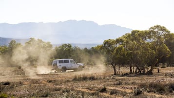 En vit SUV kör genom ett torrt, öppet landskap med gles vegetation och ett bergskedja i bakgrunden.