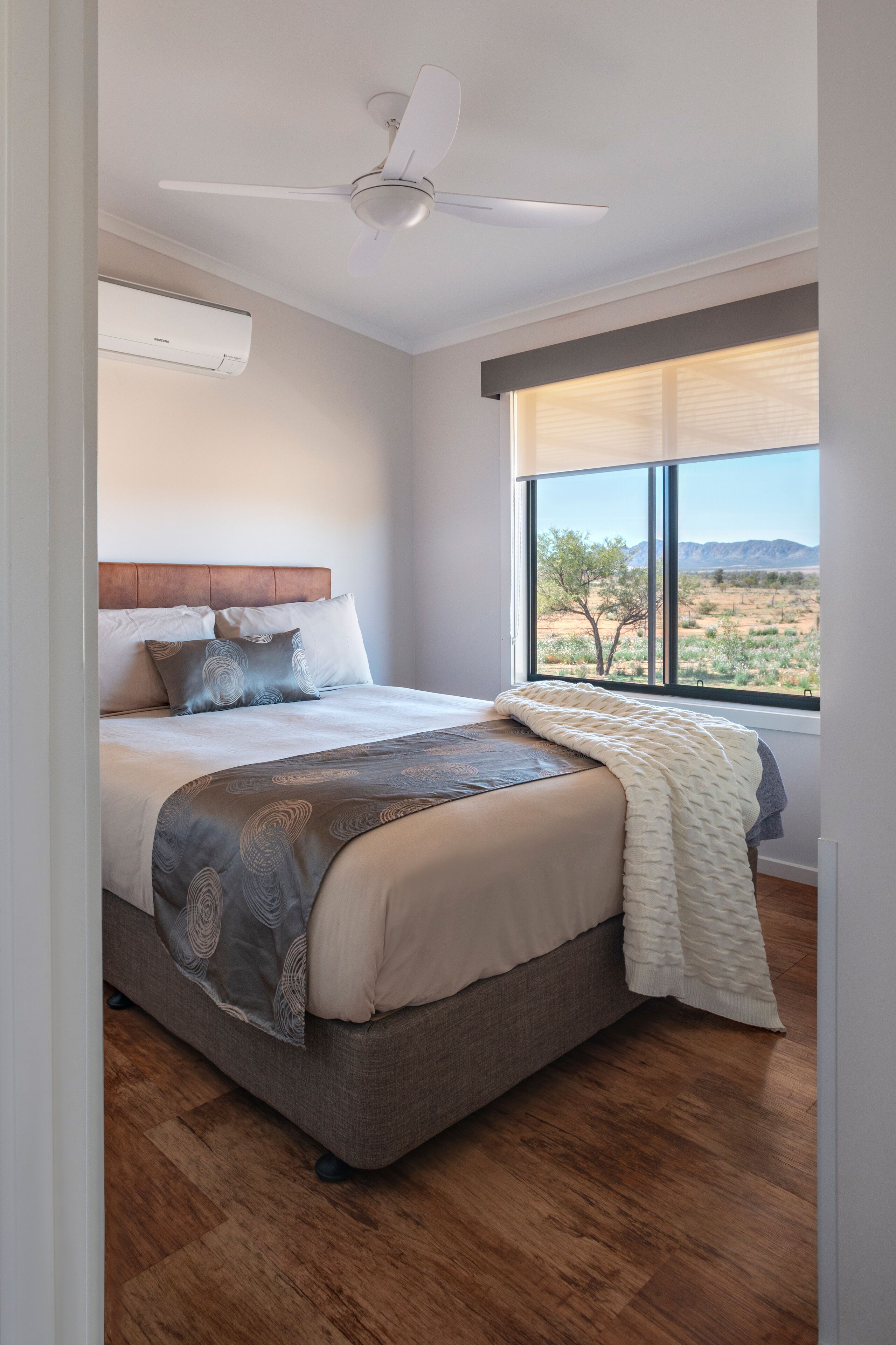 A bedroom with a bed, a ceiling fan, a window with a view of the desert, and an air conditioning unit.