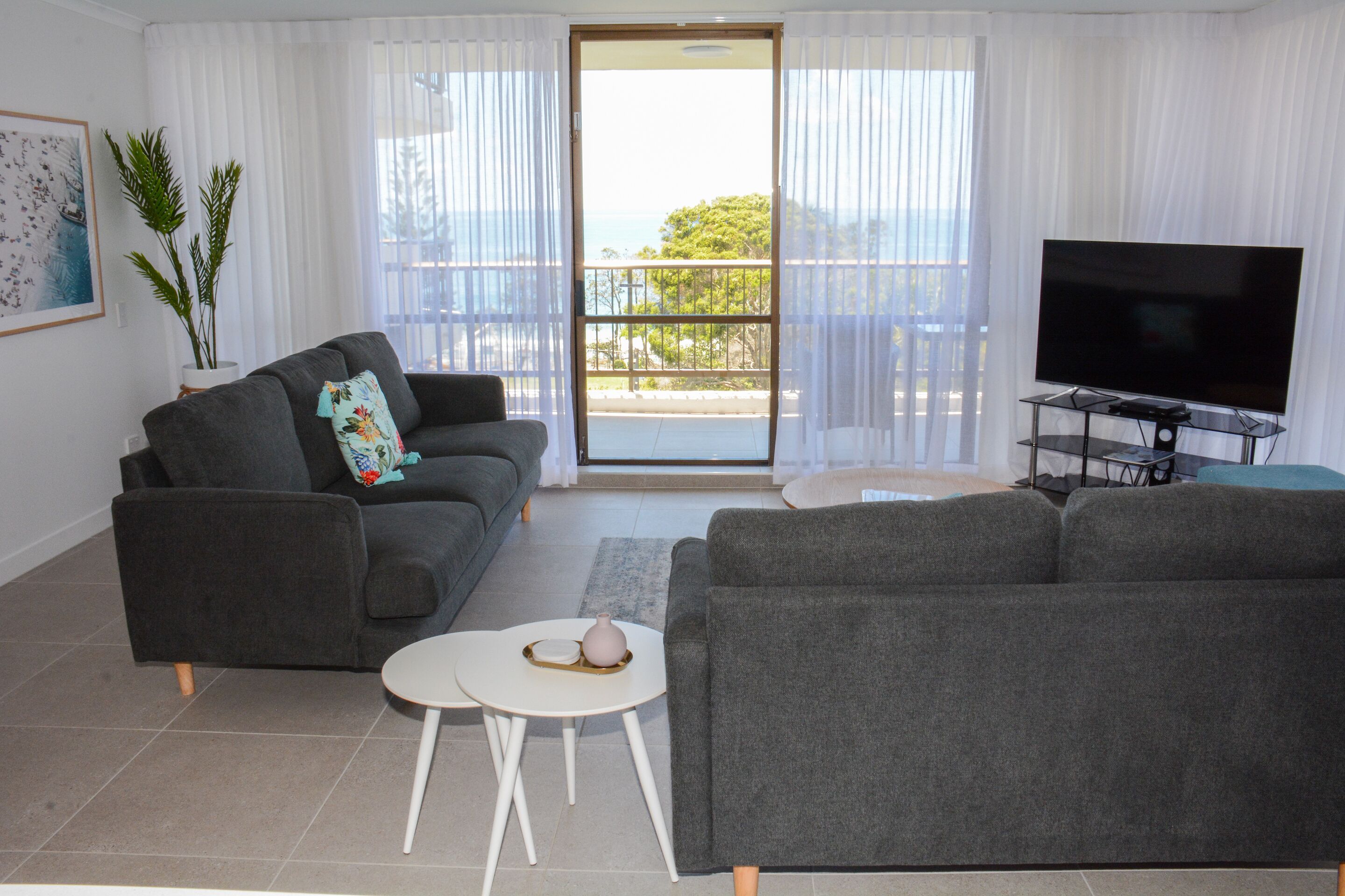 A living room with two grey sofas, a small white table, a TV on a stand, and a balcony with a view.