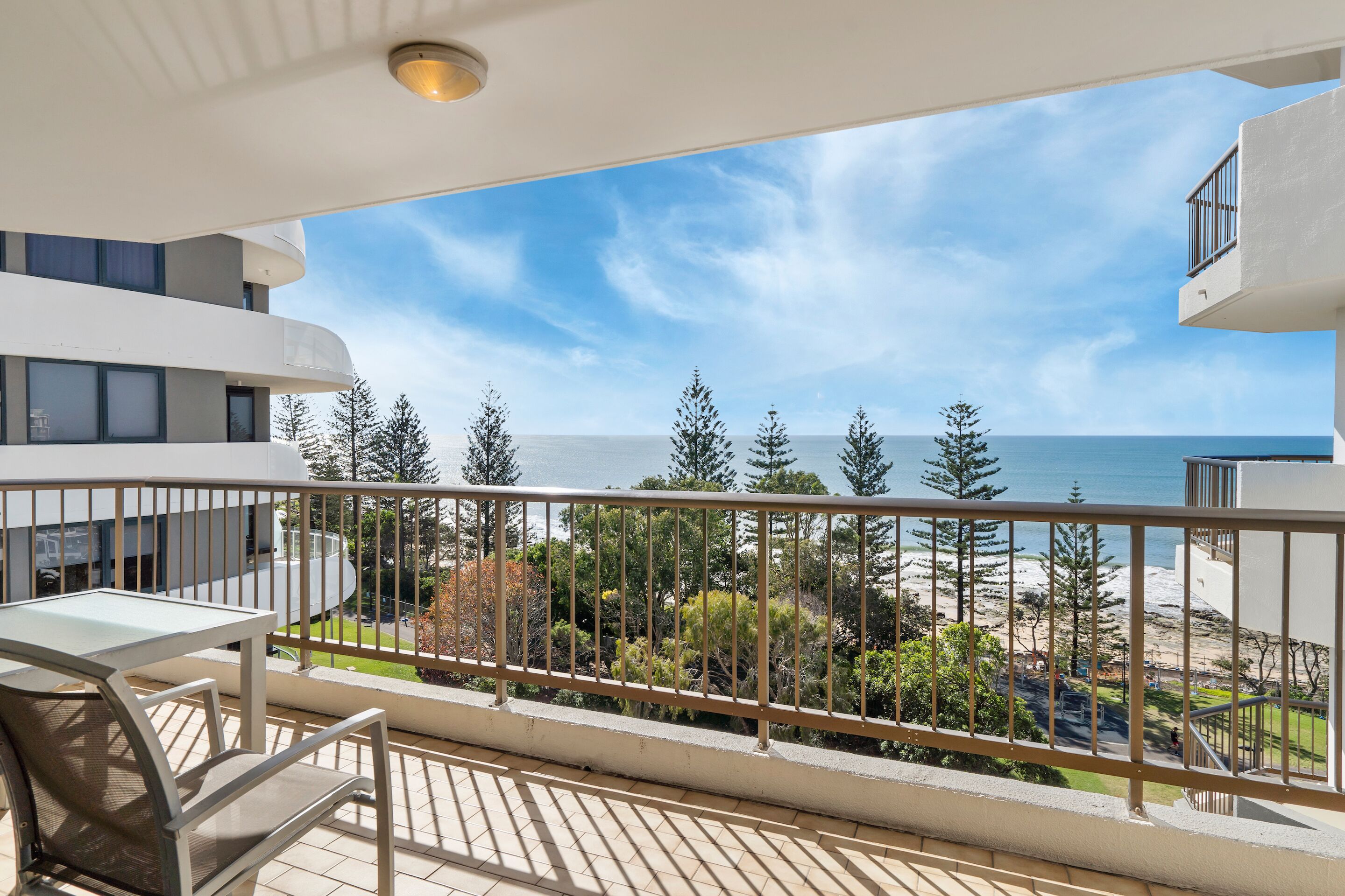 A balcony with a view of the ocean and trees, a table, and chairs.