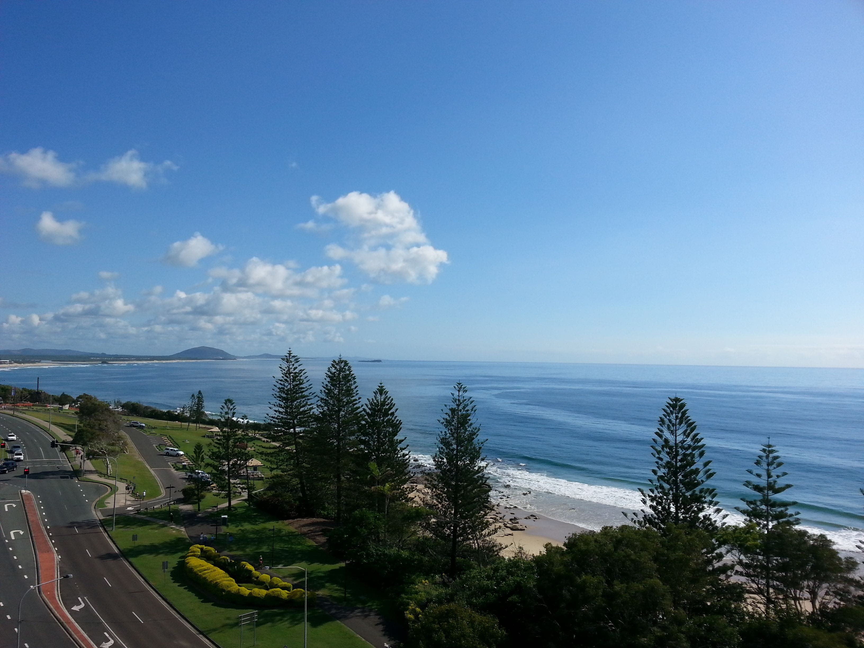 A coastal road with a beach and ocean view, surrounded by greenery and trees.