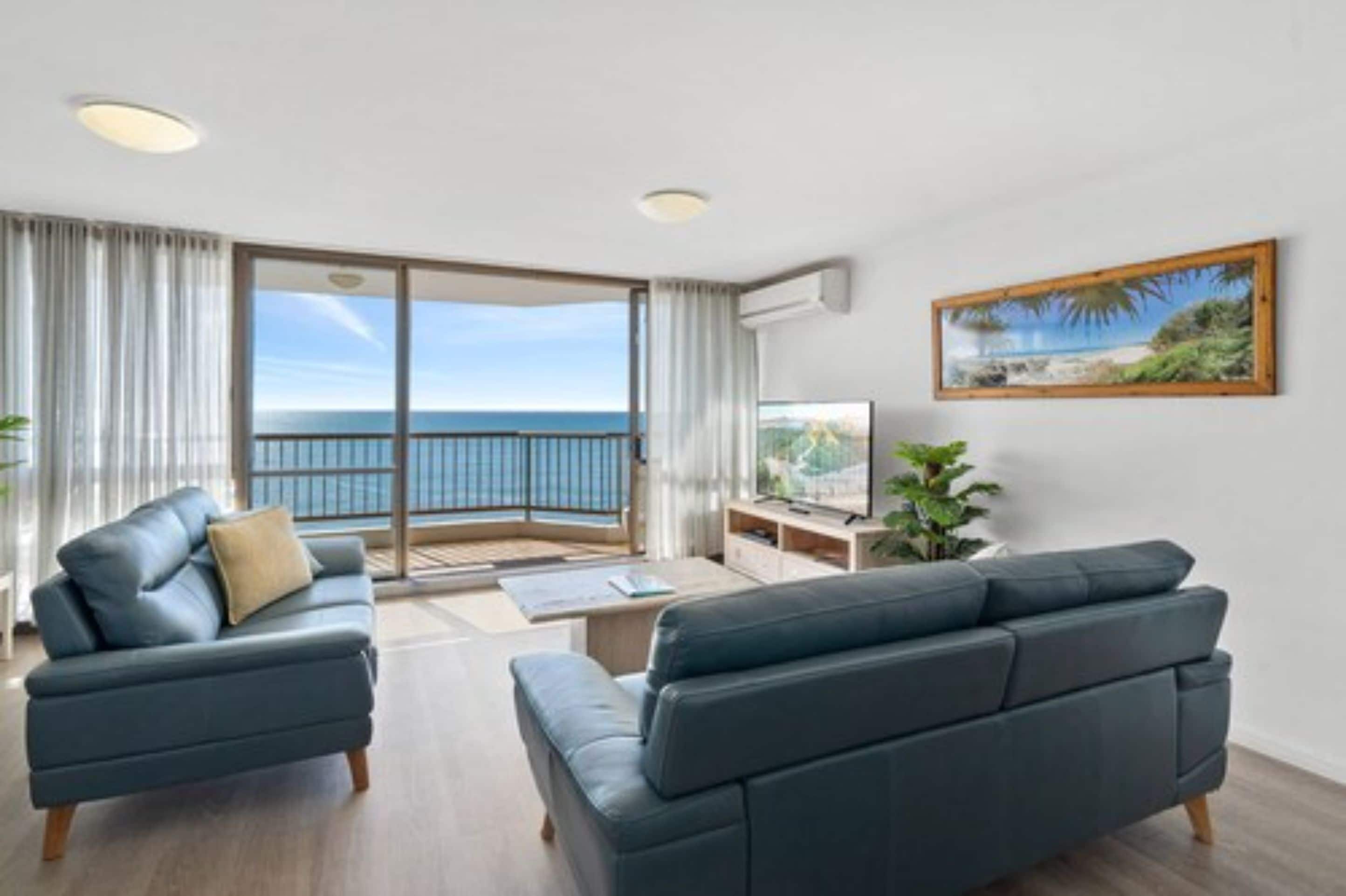 Una sala de estar moderna con un gran ventanal corredero que da a un balcón con vista al mar. Se observa un sofá, una mesa de centro y un televisor sobre un mueble.