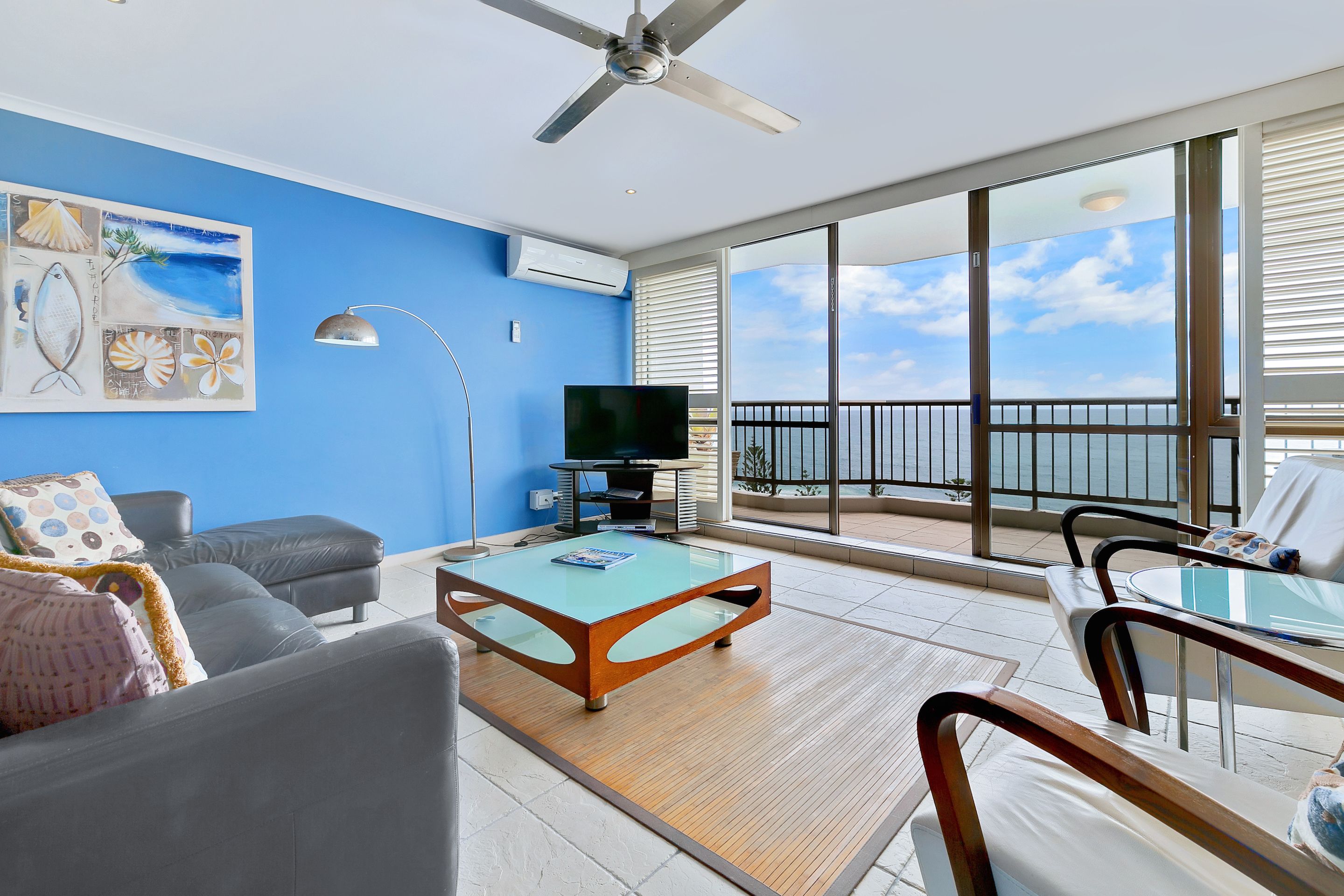 A modern living room with a glass coffee table, a sofa, and a TV. There is a balcony with a view of the ocean.