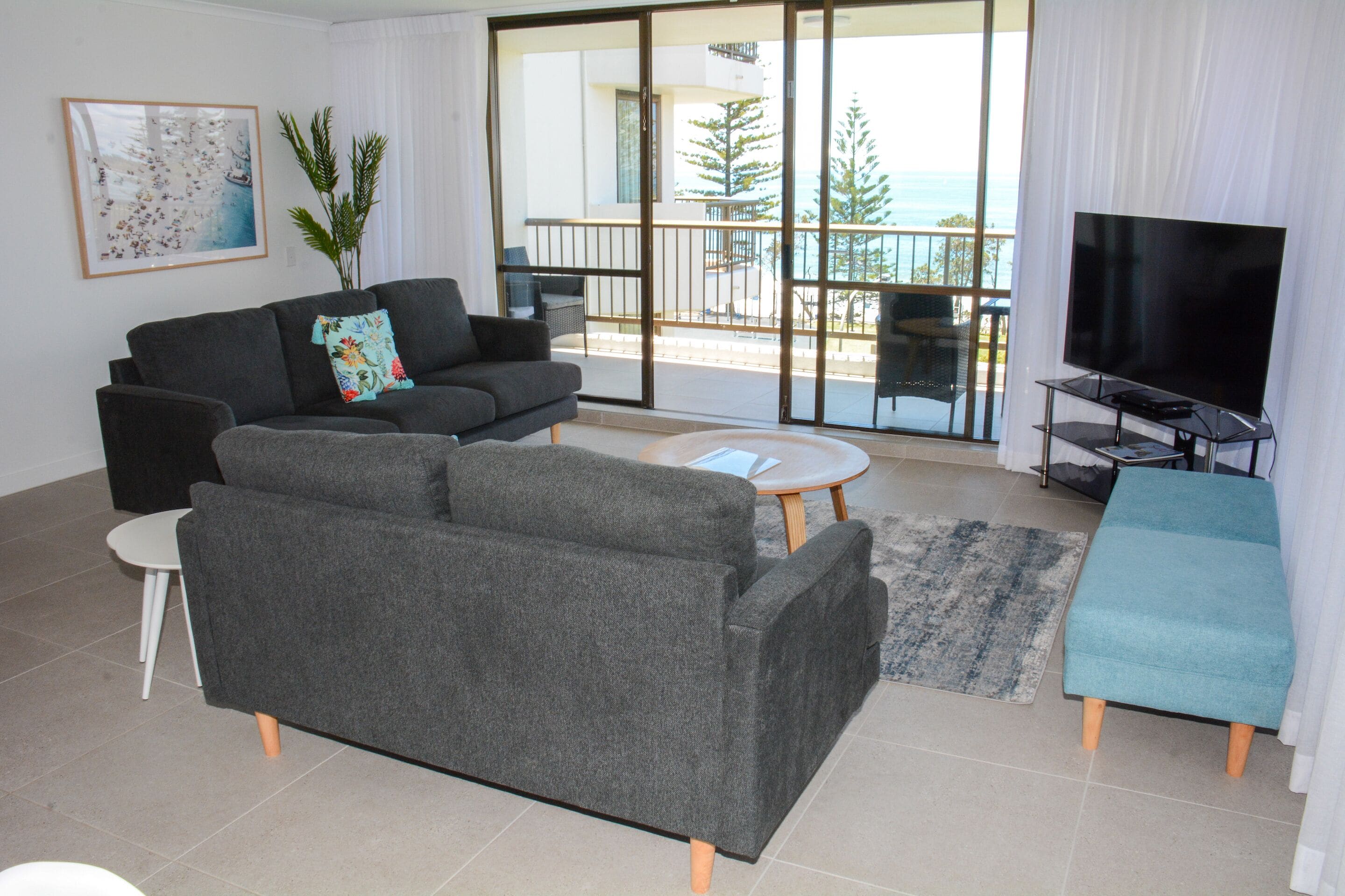 A modern living room with a grey sofa, a television on a stand, and a balcony with a view of the sea.