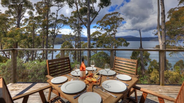 A wooden deck with a table set for dining, overlooking a lake with trees and mountains in the background. - Stewarts Bay Lodge (Port Arthur)