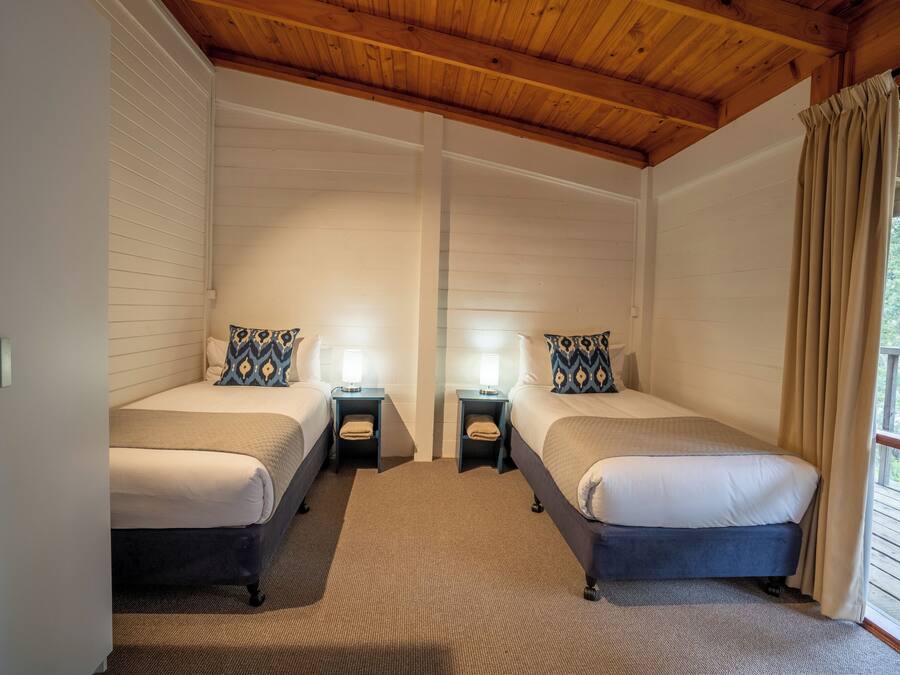 Une chambre avec deux lits, chacun doté d’une table de chevet et d’une lampe, des poutres apparentes en bois au plafond et une fenêtre avec des rideaux.