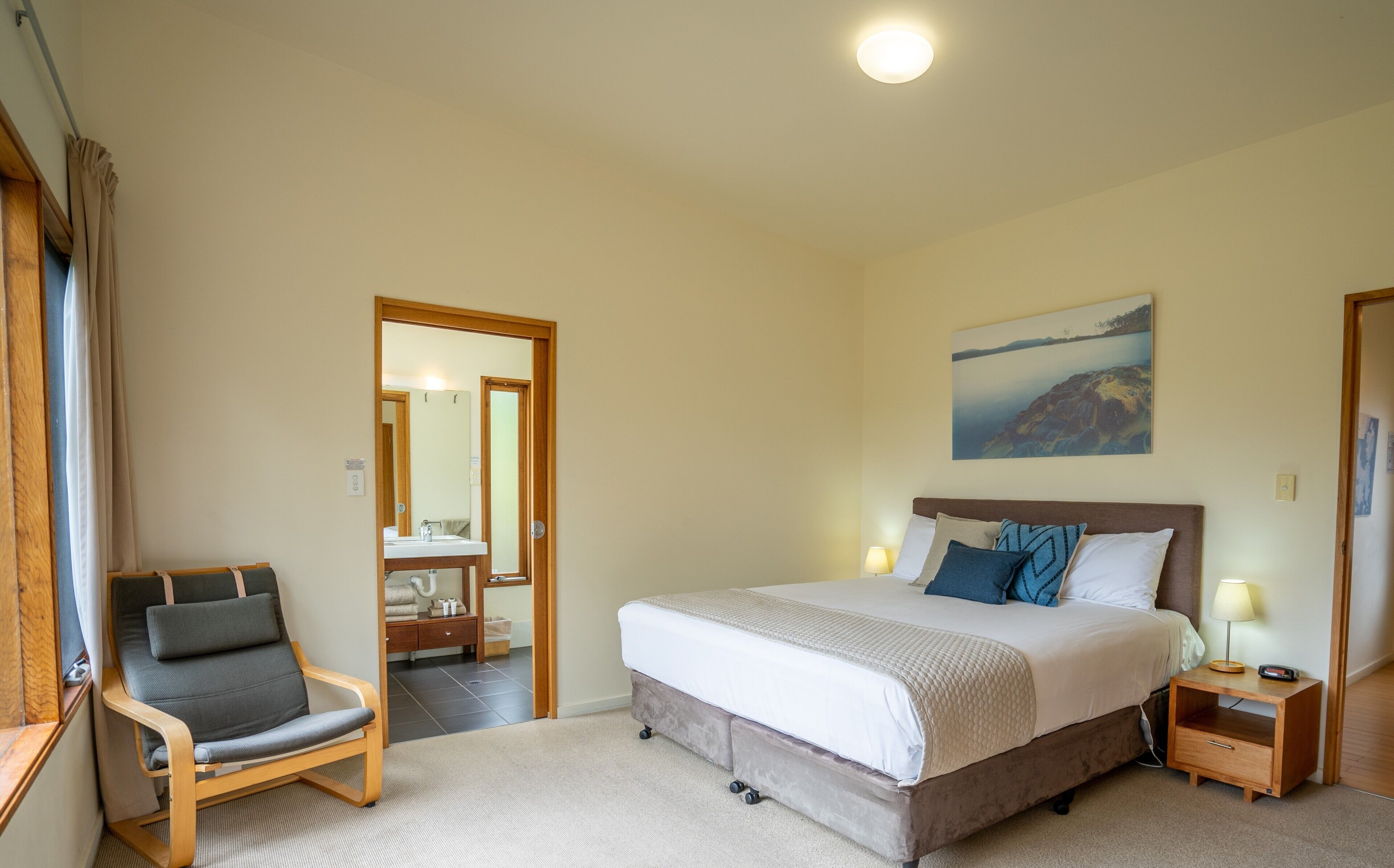 A bedroom with a bed, a chair, a bedside table, a lamp, and a painting on the wall.