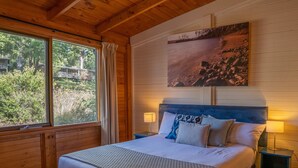 A bedroom with a large bed, two bedside tables, a window with curtains, a painting on the wall, and a view of greenery outside. - Stewarts Bay Lodge (Port Arthur)