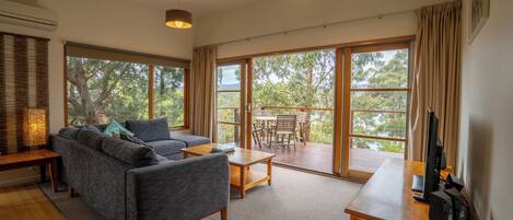 A modern living room with a sofa, coffee table, and a desk with a computer. Large windows offer a view of the outdoors.