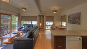 A modern living room with a sofa, dining table, and chairs. There is a kitchen area with a refrigerator and cabinets. Large windows provide views of the outdoors. - Stewarts Bay Lodge (Port Arthur)