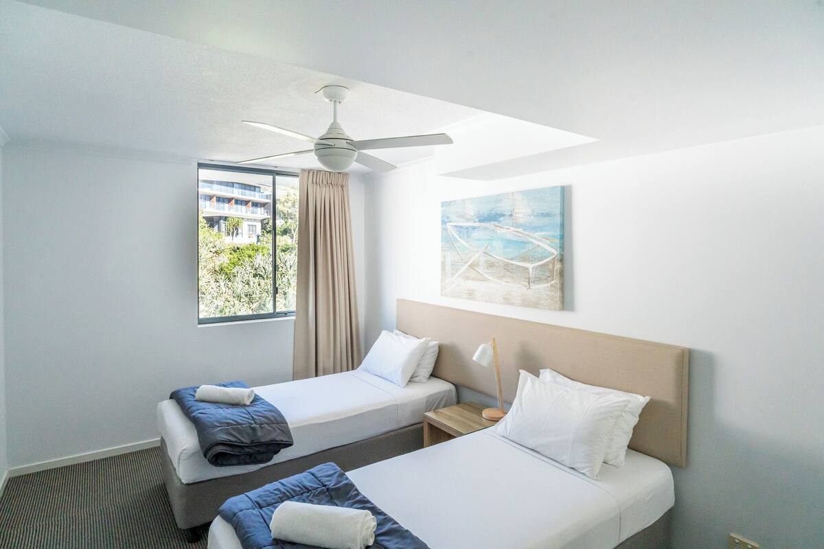 A hotel room with two beds, a ceiling fan, a window with curtains, and a painting on the wall.