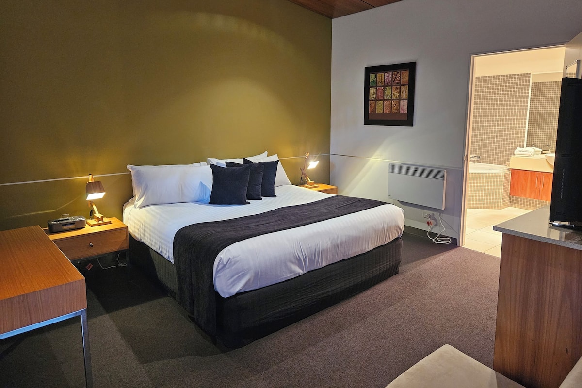 Una habitación de hotel con una cama grande, dos mesitas de noche con lámparas, una obra de arte enmarcada en la pared y un baño visible a través de una puerta abierta.