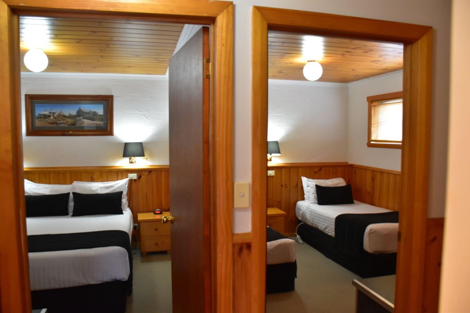 A room with two beds, a wooden headboard, a nightstand with a lamp, and a framed picture on the wall.