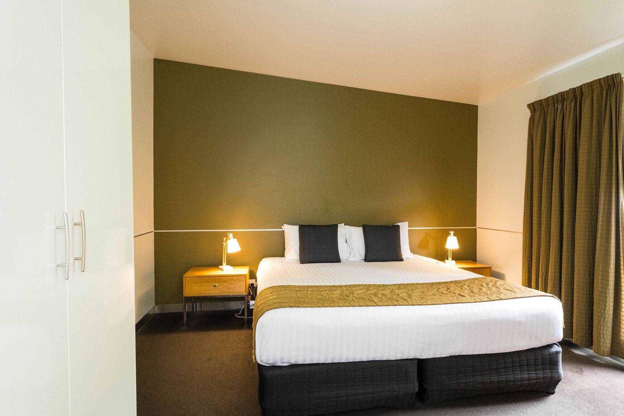 A hotel room with a large bed, two bedside tables with lamps, a wardrobe, and a window with curtains.