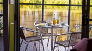 Una mesa de comedor con sillas en una terraza, con vista a un cuerpo de agua y follaje.