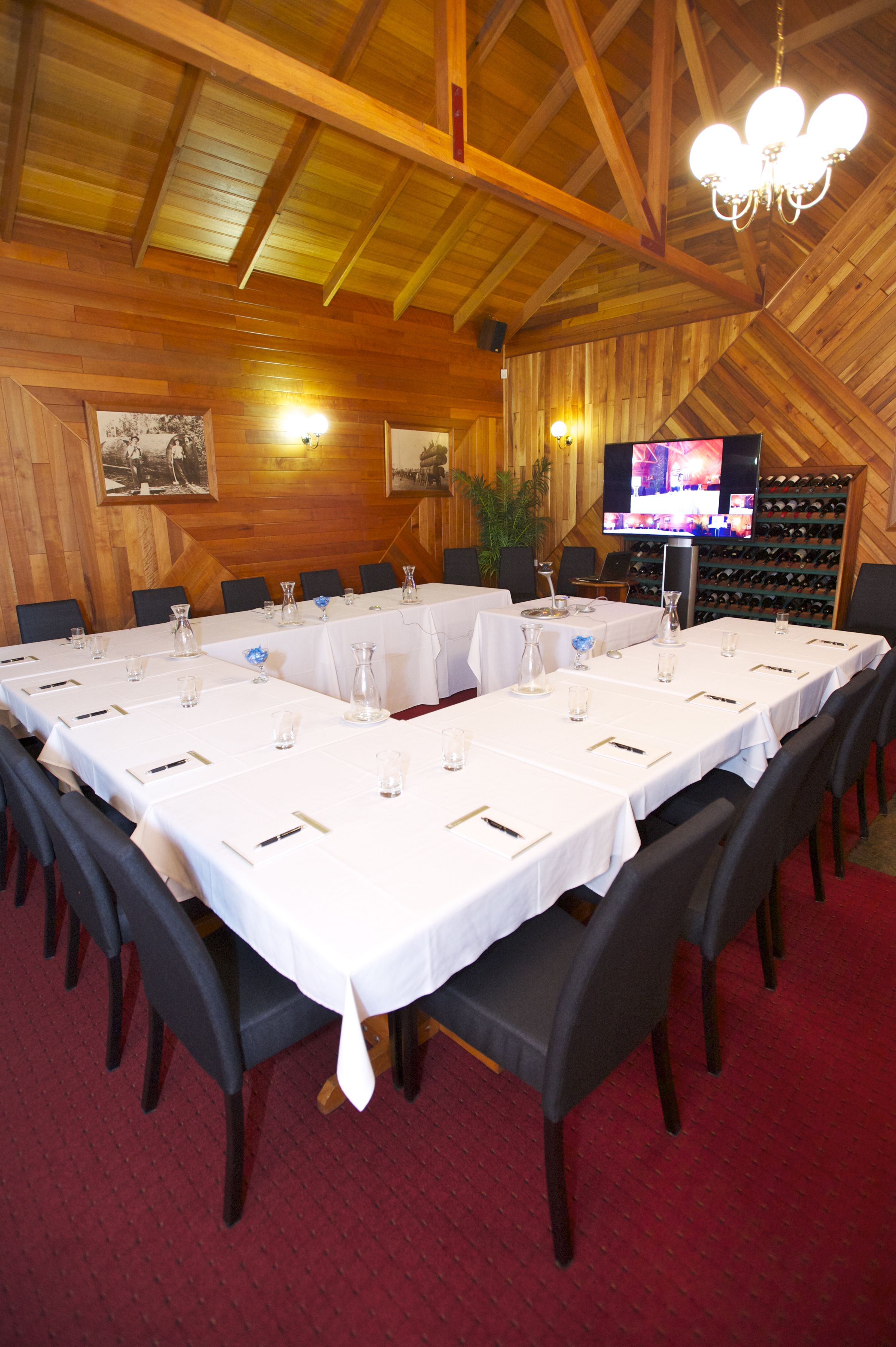 Un salón de conferencias con largas mesas dispuestas para una reunión, paredes de madera, una araña y una estantería para vinos al fondo.