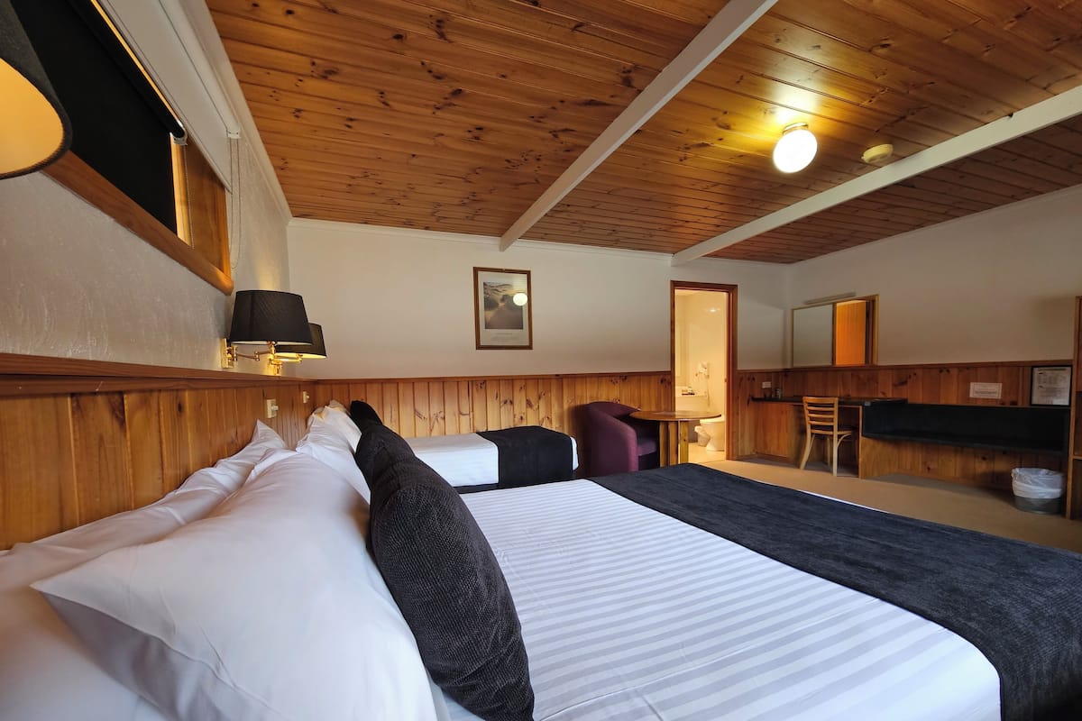 Habitación de hotel con techo de madera, cama con ropa de cama blanca y negra, una mesita, una silla y un baño visible a través de una puerta abierta.