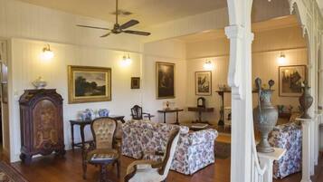 Un salon spacieux avec un plafond haut, un ventilateur de plafond et une cheminée. On y trouve plusieurs meubles, dont un canapé, des fauteuils et une table. Les murs sont ornés de tableaux encadrés et une grande vasque est posée sur une table d’appoint.