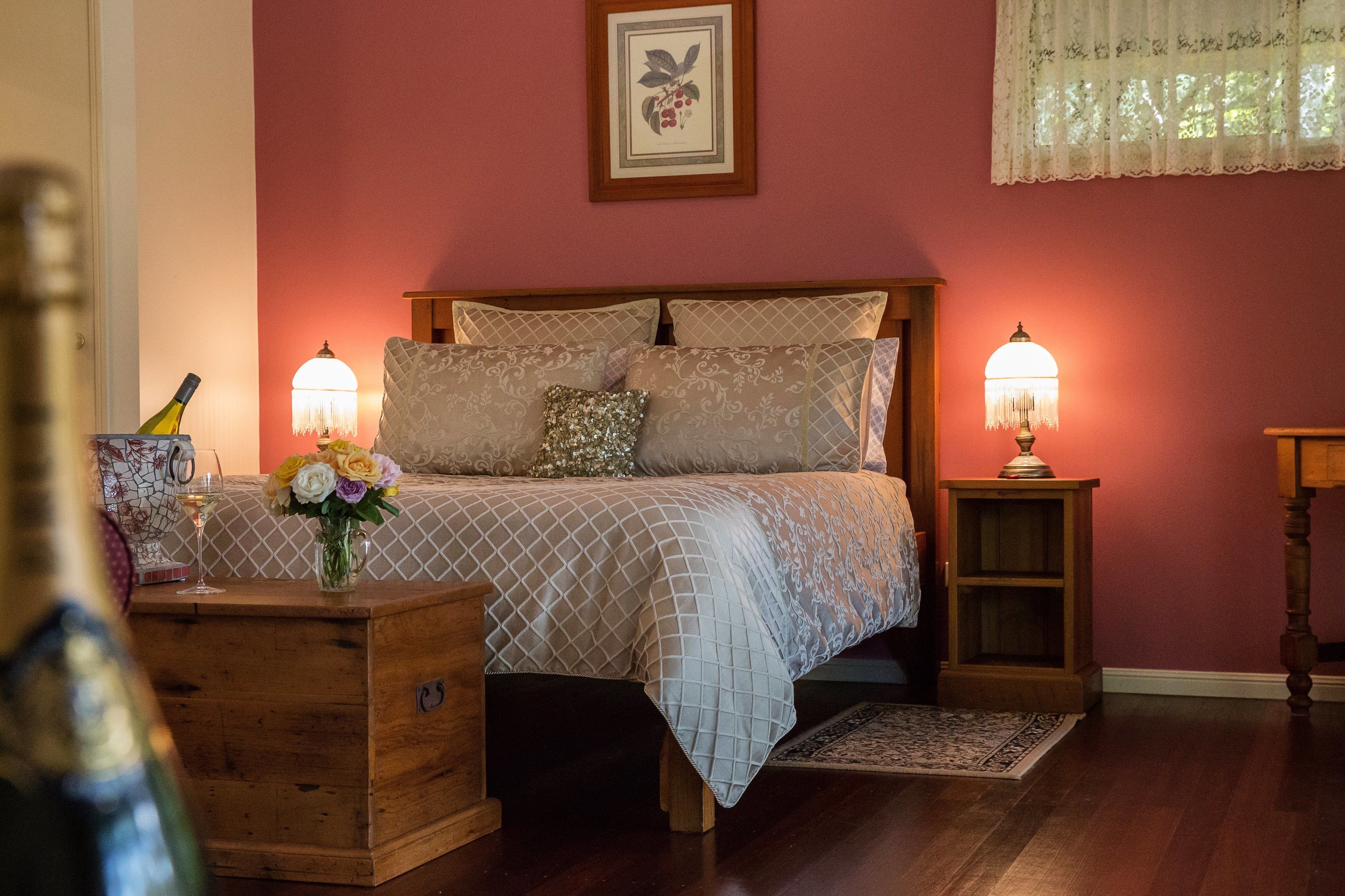 A bedroom with a wooden bed, two bedside lamps, a nightstand with a bottle and flowers, and a framed picture on the wall.