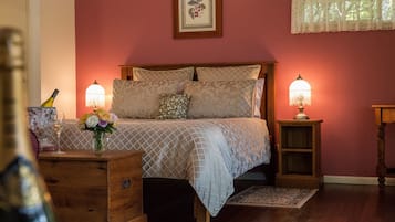 A bedroom with a wooden bed, two bedside lamps, a nightstand with a bottle and flowers, and a framed picture on the wall.