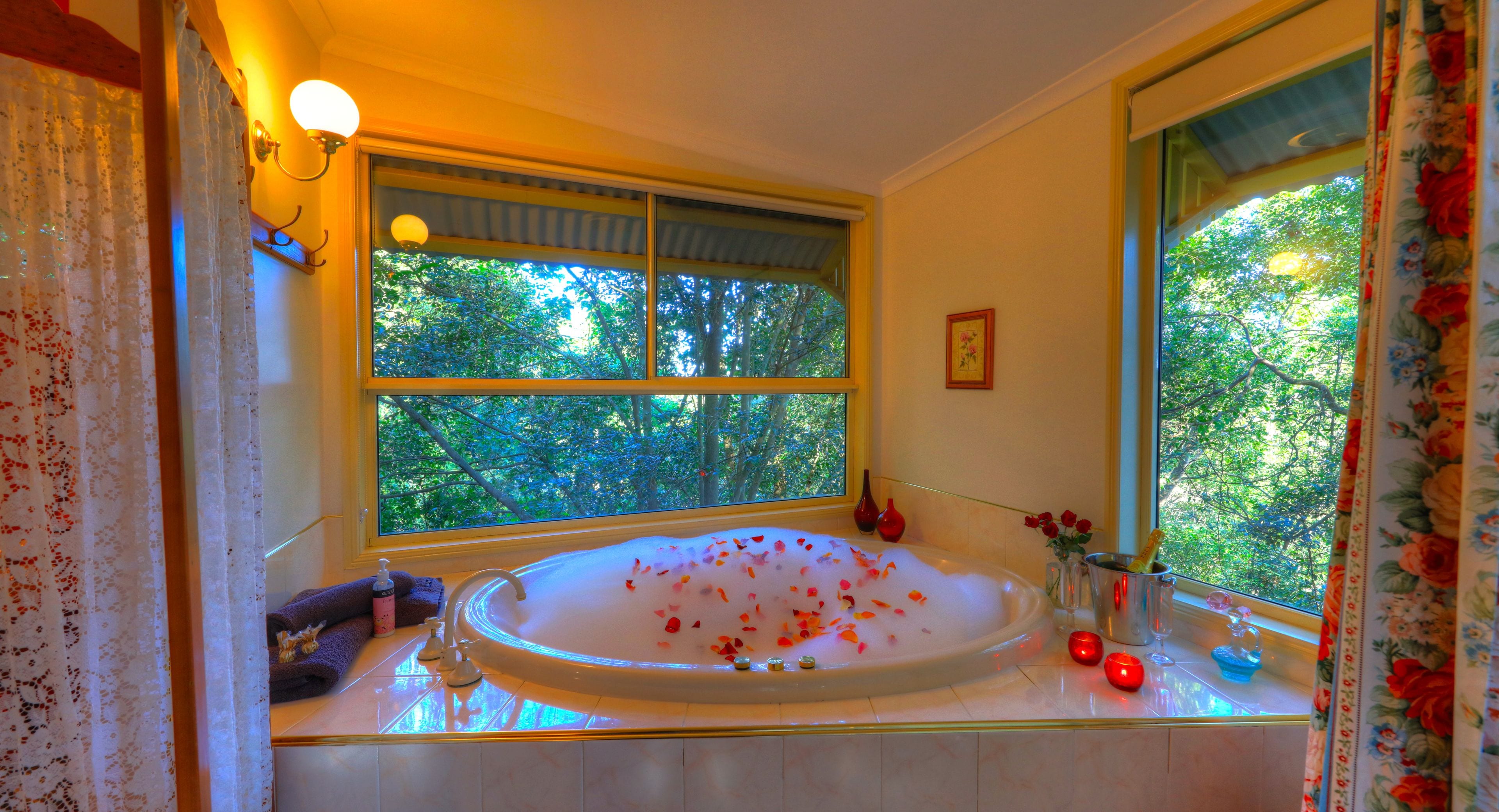 A bathroom with a large bathtub filled with water and rose petals, a window with a view of trees, and a vase with flowers on the counter.