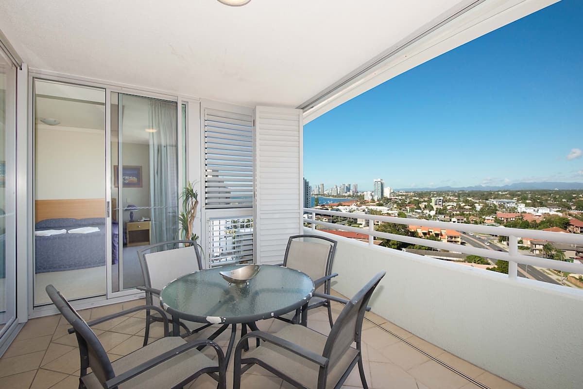 Ein Balkon mit einem Glastisch und Stühlen, ein Bett mit blauer Kopfteil und Blick auf die Stadt.