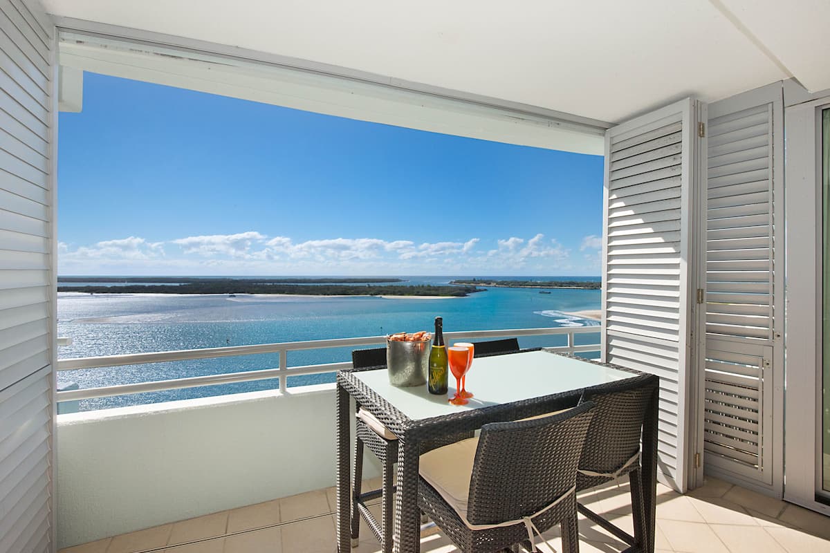 Ein Balkon mit Tisch und Stühlen, der Blick auf das klare, blaue Meer und entferntes Land freigibt.