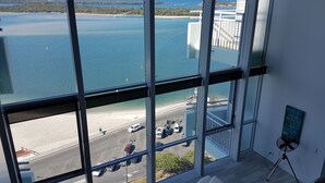 Quarto com amplas janelas com vista para uma praia e um estacionamento.