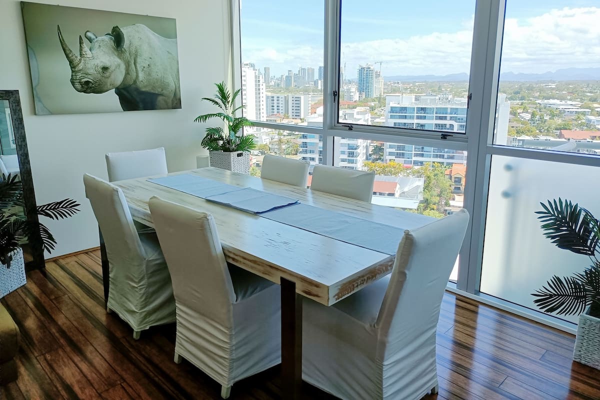 Ein moderner Essbereich mit großem Tisch, weißen Stühlen und Blick auf die Stadt durch hohe Fenster.