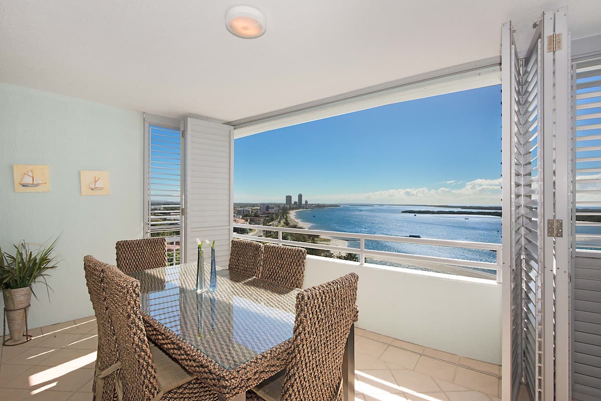 Ein Balkon mit Glastisch und Korbstühlen, mit Blick auf eine Küstenstadt, einen Strand und das Meer.