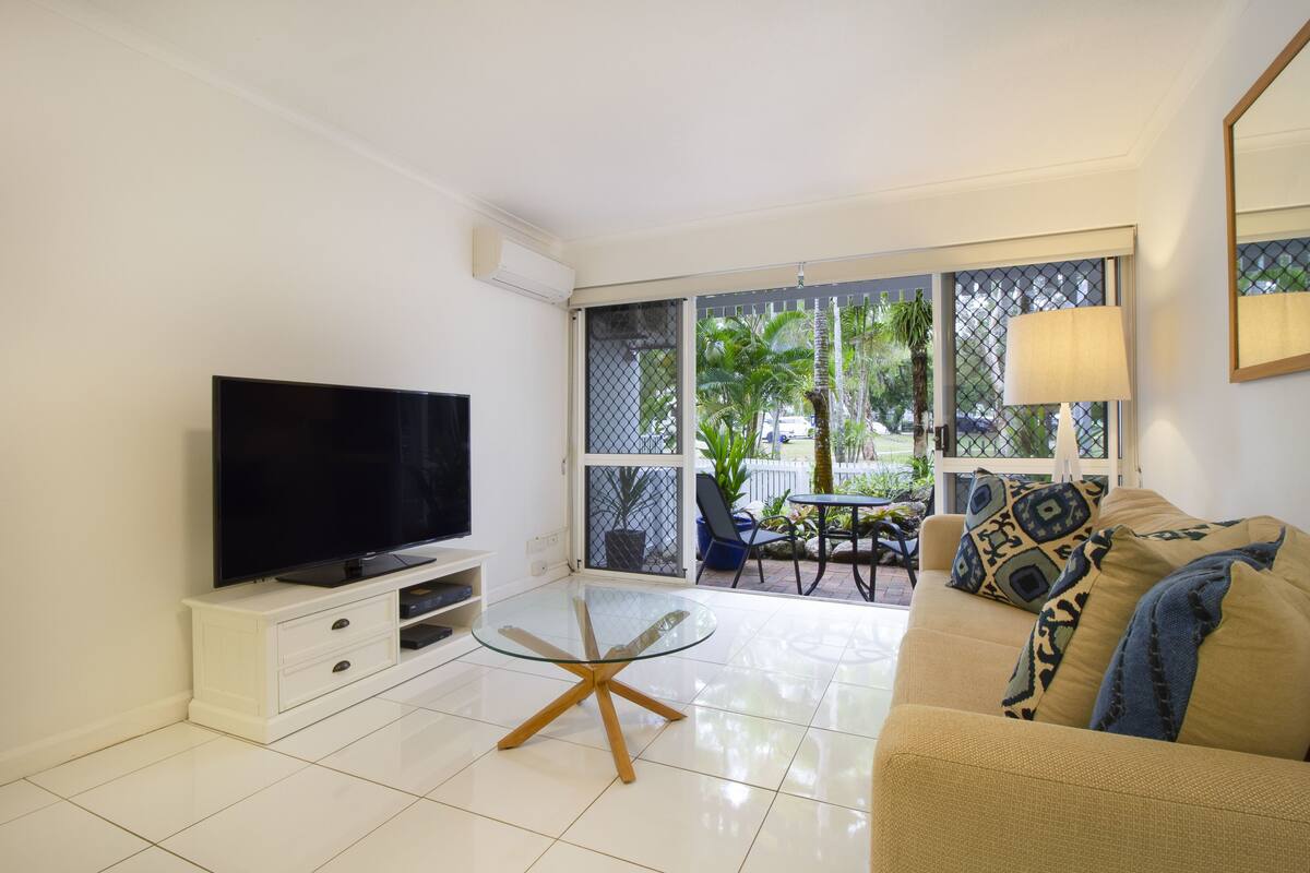 Sala de estar com televisão, mesa de centro de vidro, sofá e vista para o exterior através de grandes janelas.