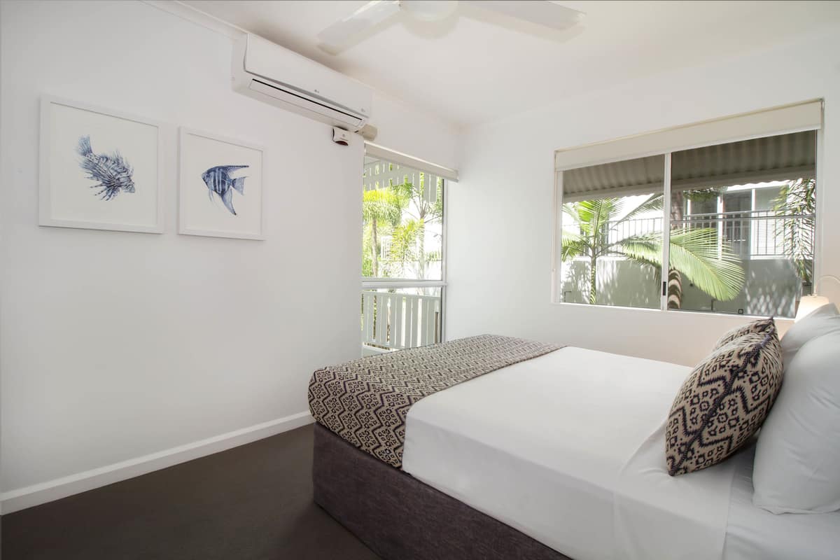 Quarto com cama, duas obras de arte com peixes emoldurados, janela com vista para palmeiras e aparelho de ar condicionado.