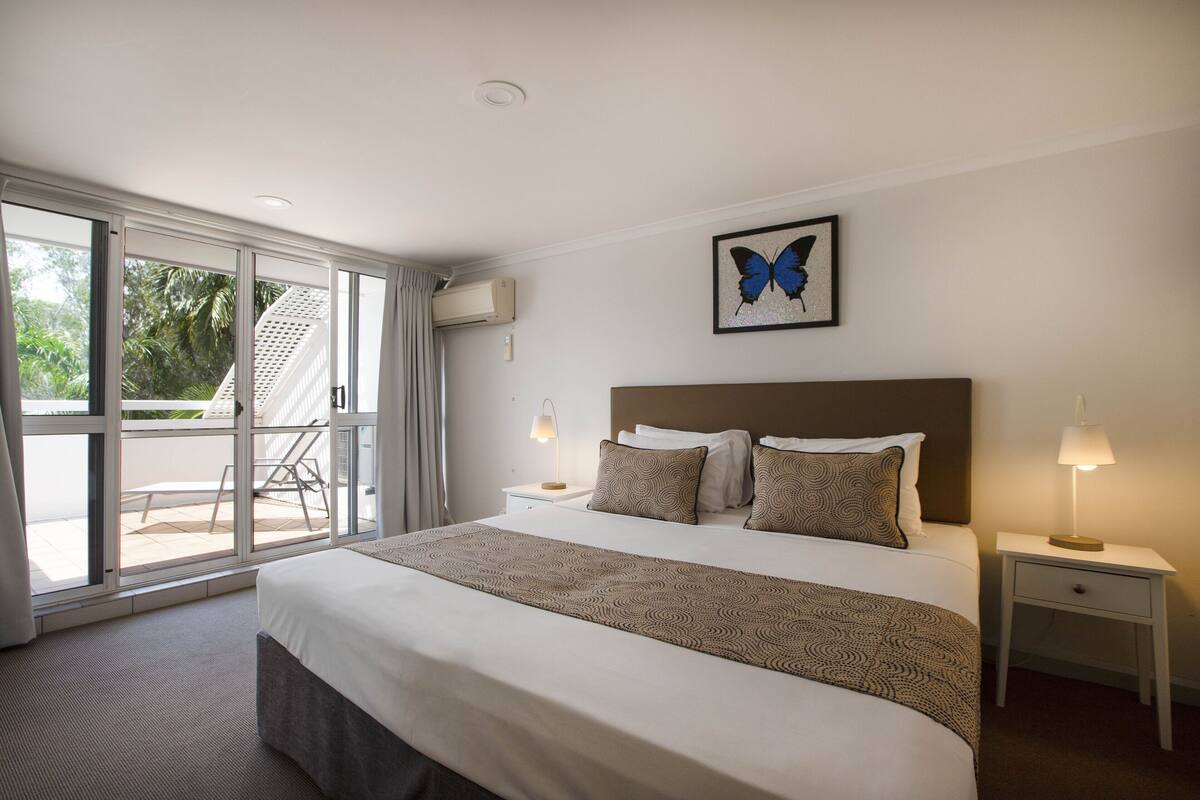 Quarto de hotel com uma cama grande, uma mesinha de cabeceira, uma luminária na parede e uma obra de arte com borboletas.