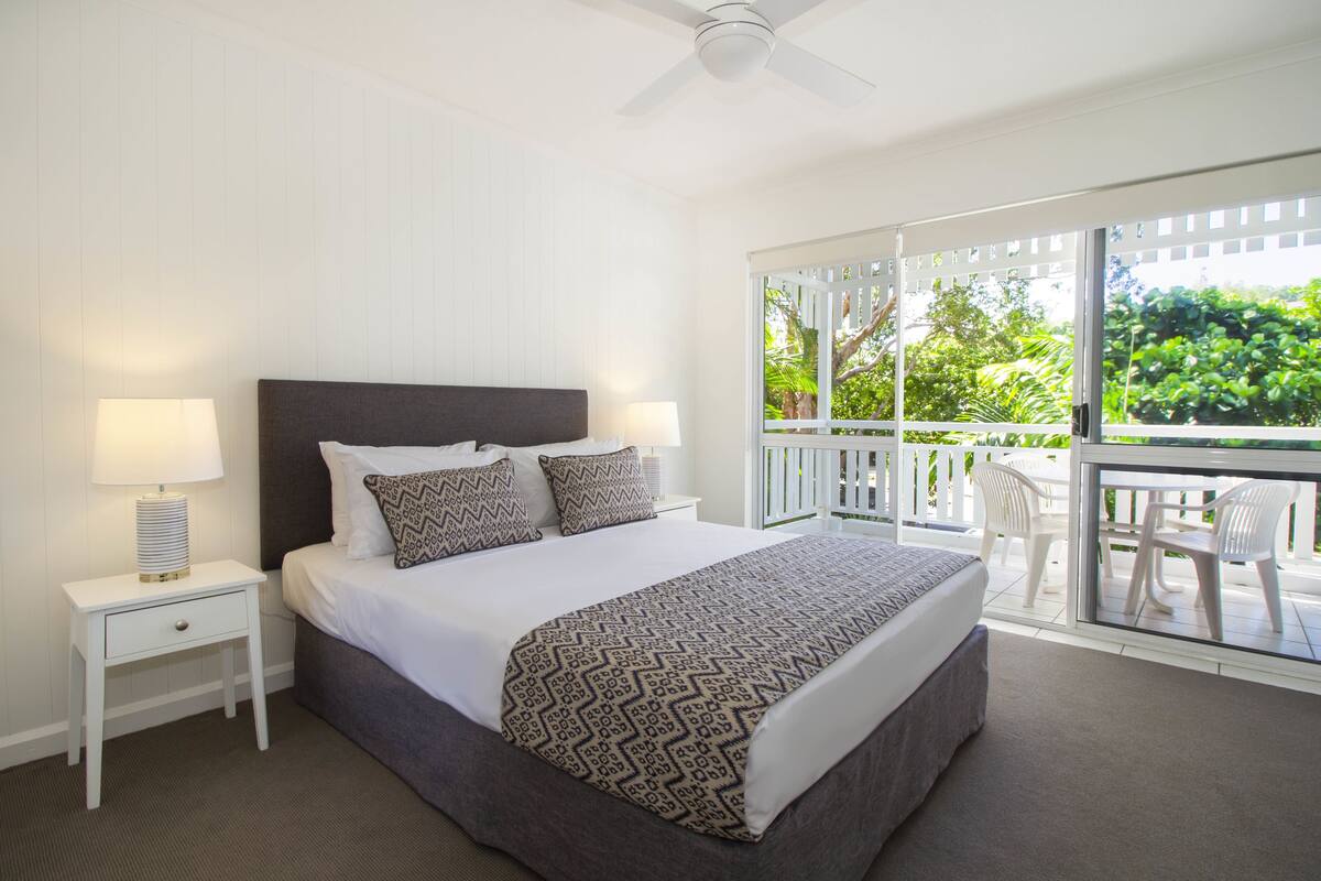 Quarto com cama, abajures, varanda com cadeiras e vista para vegetação.