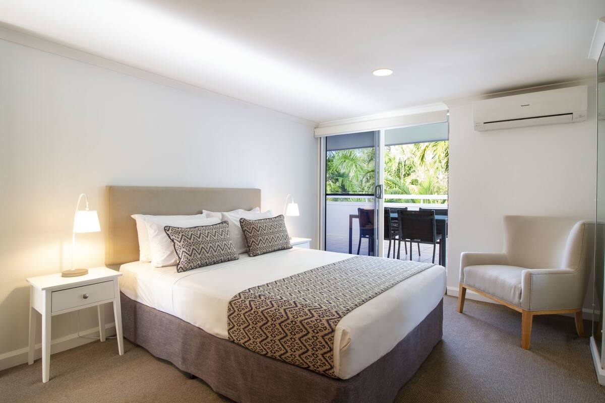 Quarto de hotel com uma cama grande, uma mesa de cabeceira com abajur, uma cadeira e vista para o exterior através de uma porta deslizante.