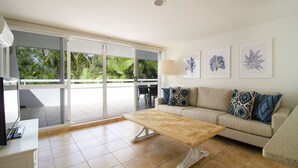 Una sala de estar moderna con un sofá, una mesa de centro y un televisor sobre un mueble. Hay ventanas amplias y un balcón con vistas a la vegetación.