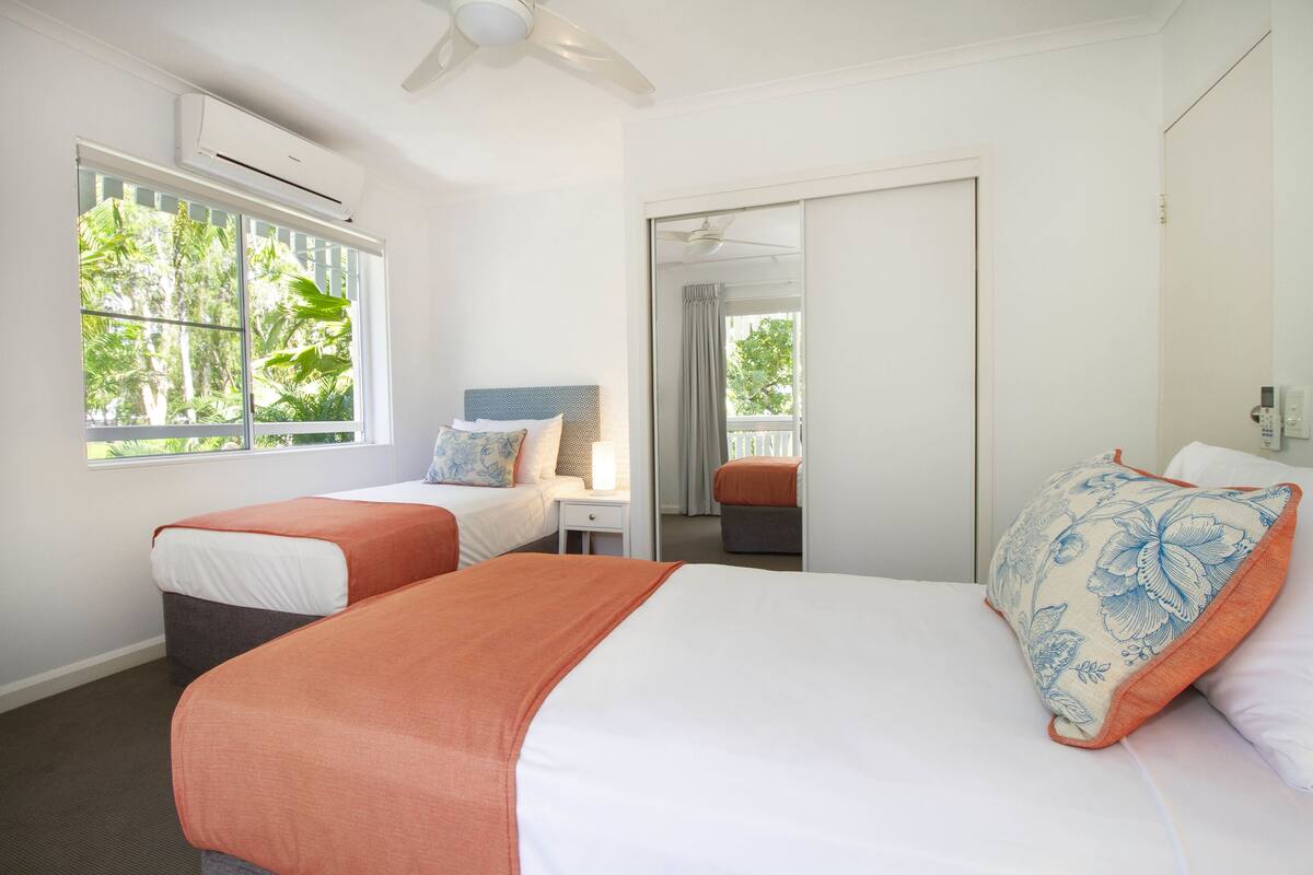 Quarto de hotel com duas camas, janela com vista para árvores, ventilador de teto e porta de correr para uma varanda.