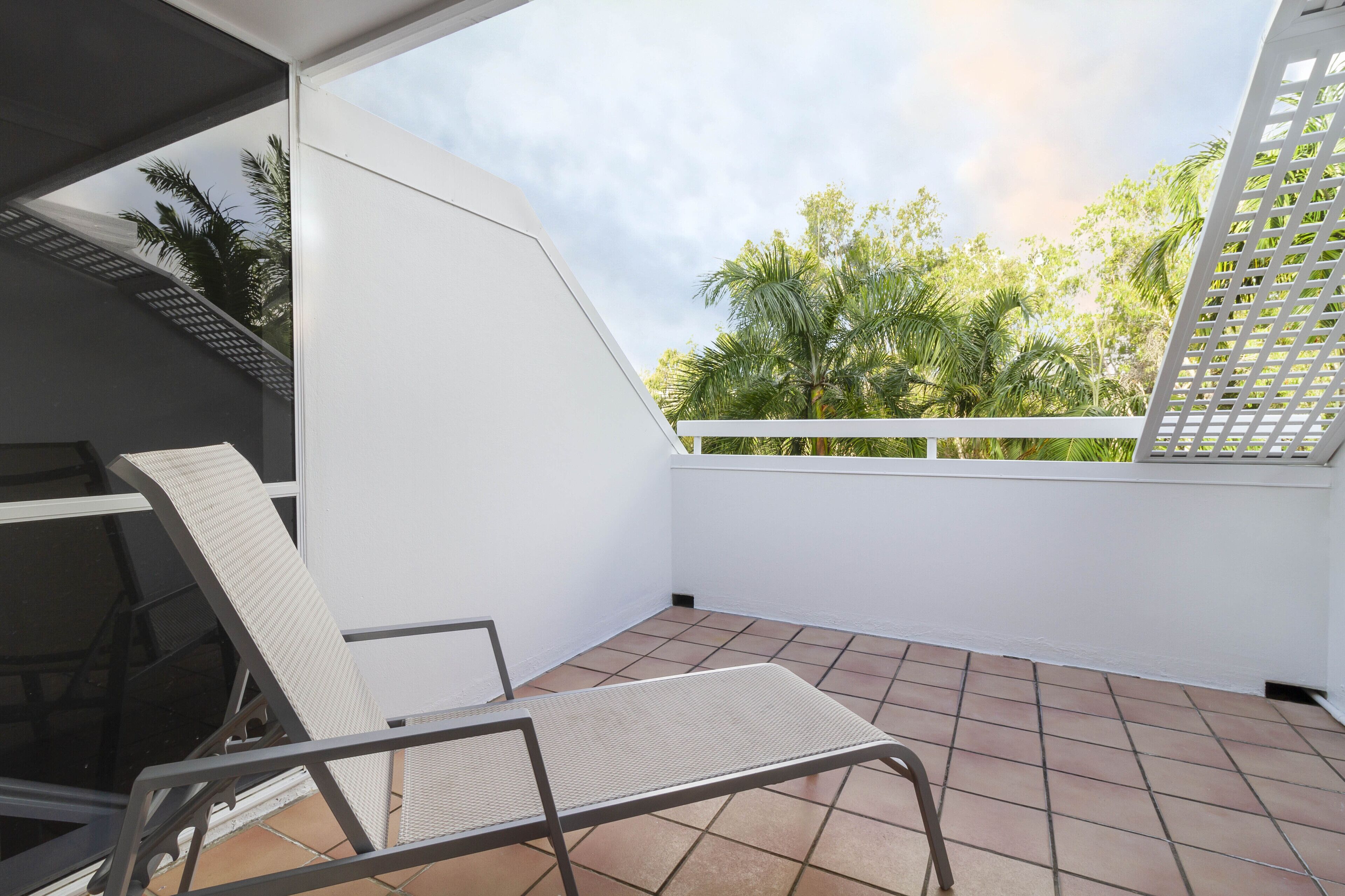 Un balcon avec une chaise blanche et une table en métal, un mur blanc et une vue sur des arbres verdoyants.