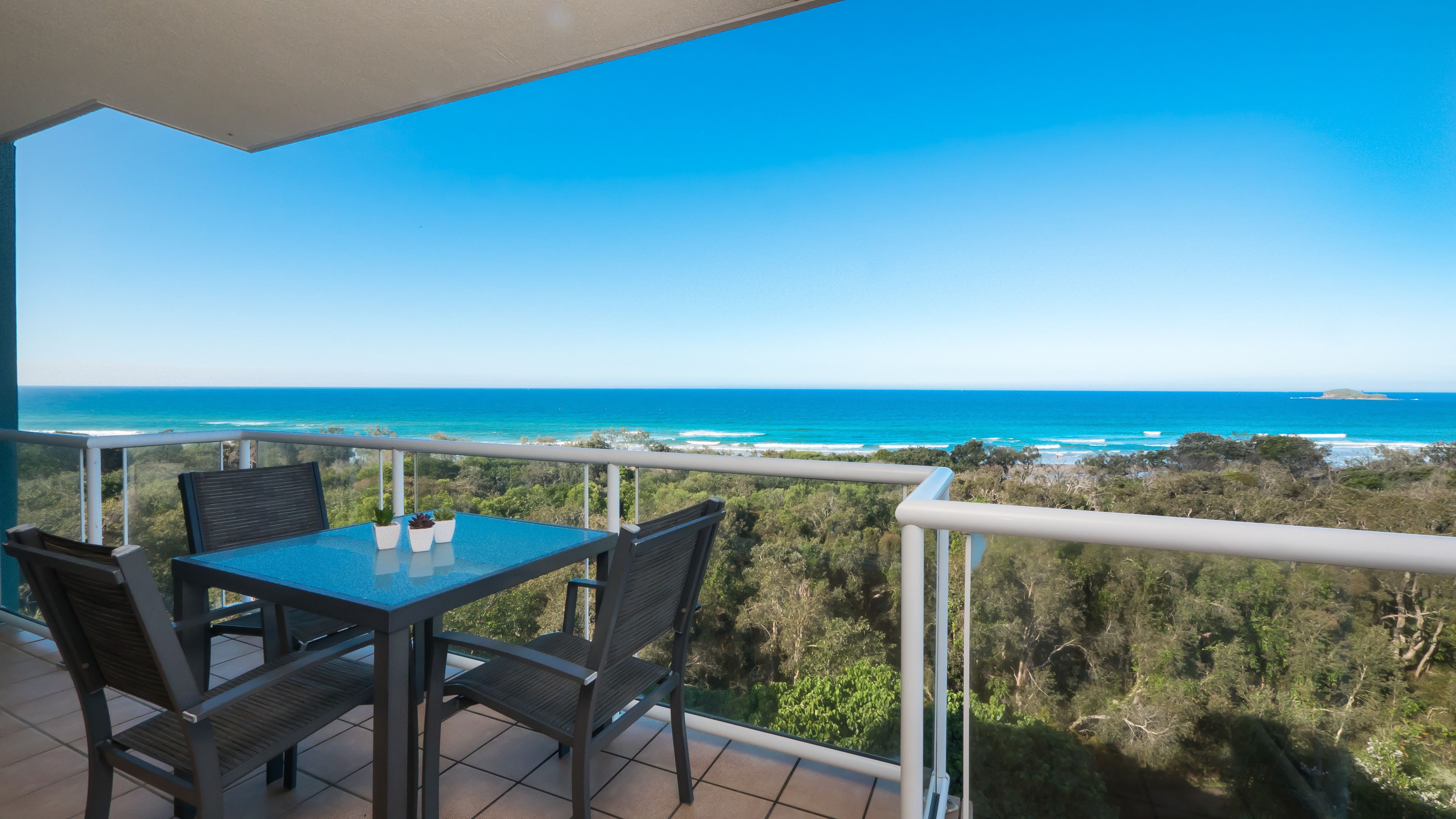 A balcony with a table and chairs, overlooking a coastal landscape with clear blue waters and distant land.