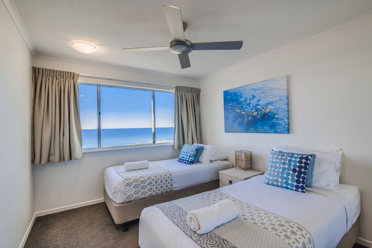 A hotel room with two beds, a ceiling fan, a painting, and a window with curtains.