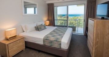 Habitación de hotel con una cama grande, televisión, cómoda de madera y balcón con vista al océano.