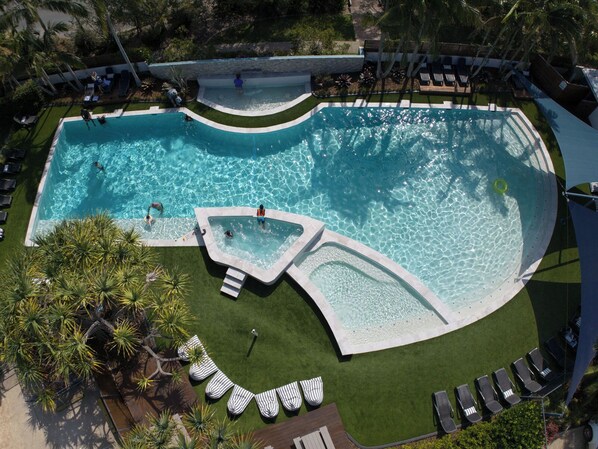 Uma piscina grande e de formato irregular, rodeada por espreguiçadeiras e palmeiras.