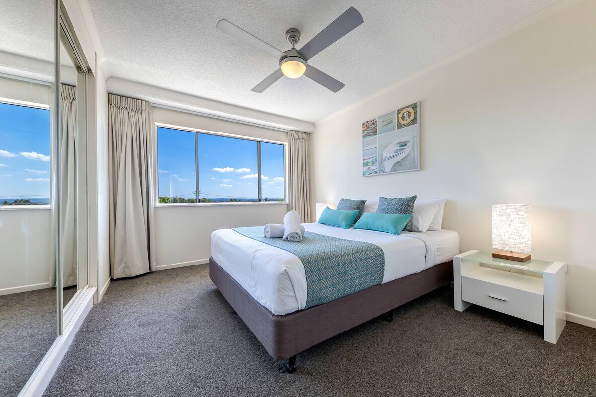A bedroom with a large bed, a nightstand, a ceiling fan, and a window with curtains.