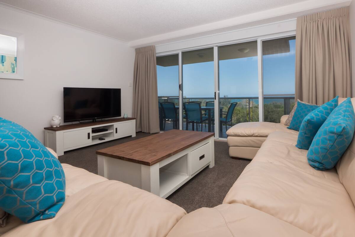 A modern living room with a sofa, a coffee table, a TV, and a balcony with outdoor furniture.