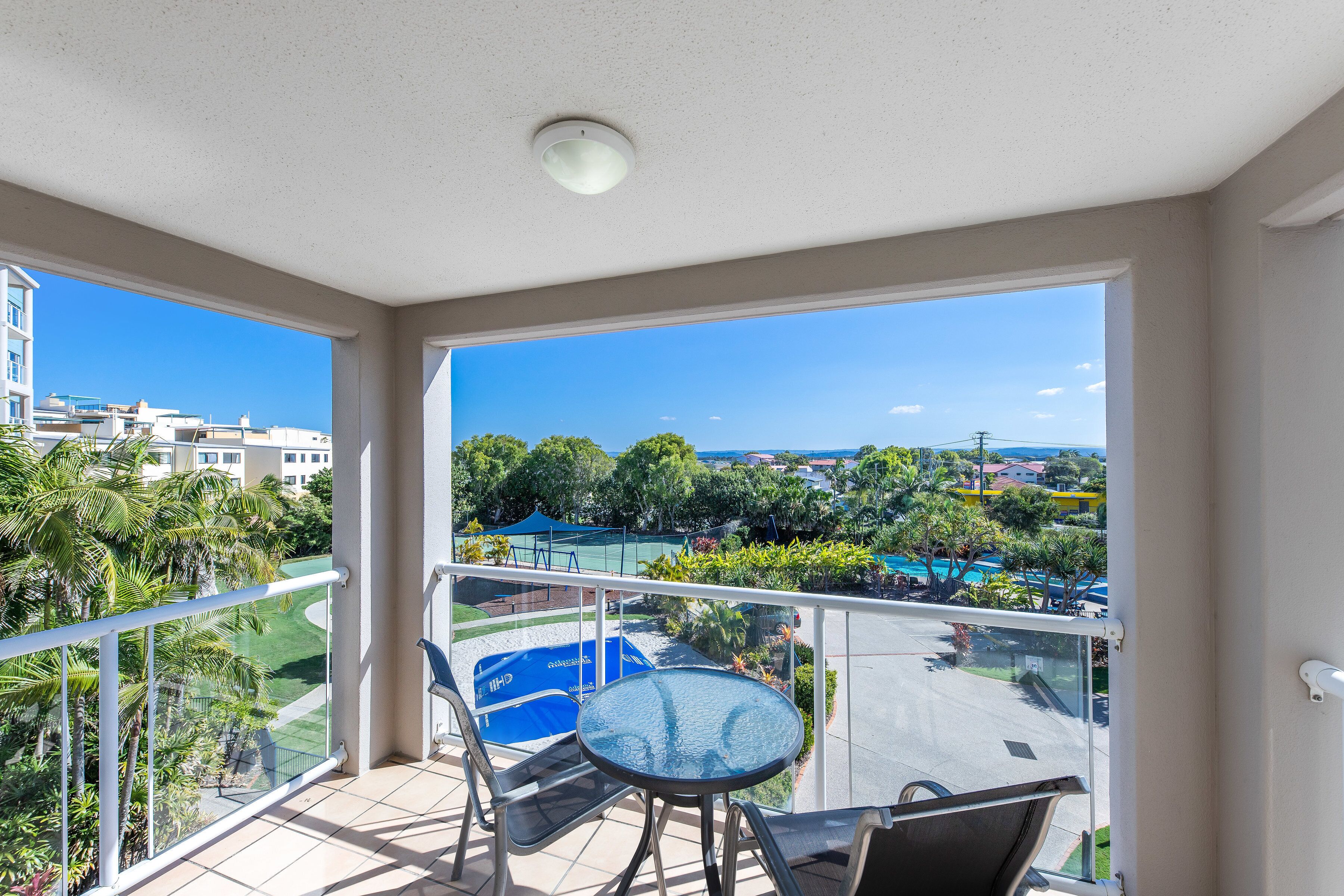 A balcony with a glass table and chairs, overlooking a pool and greenery.
