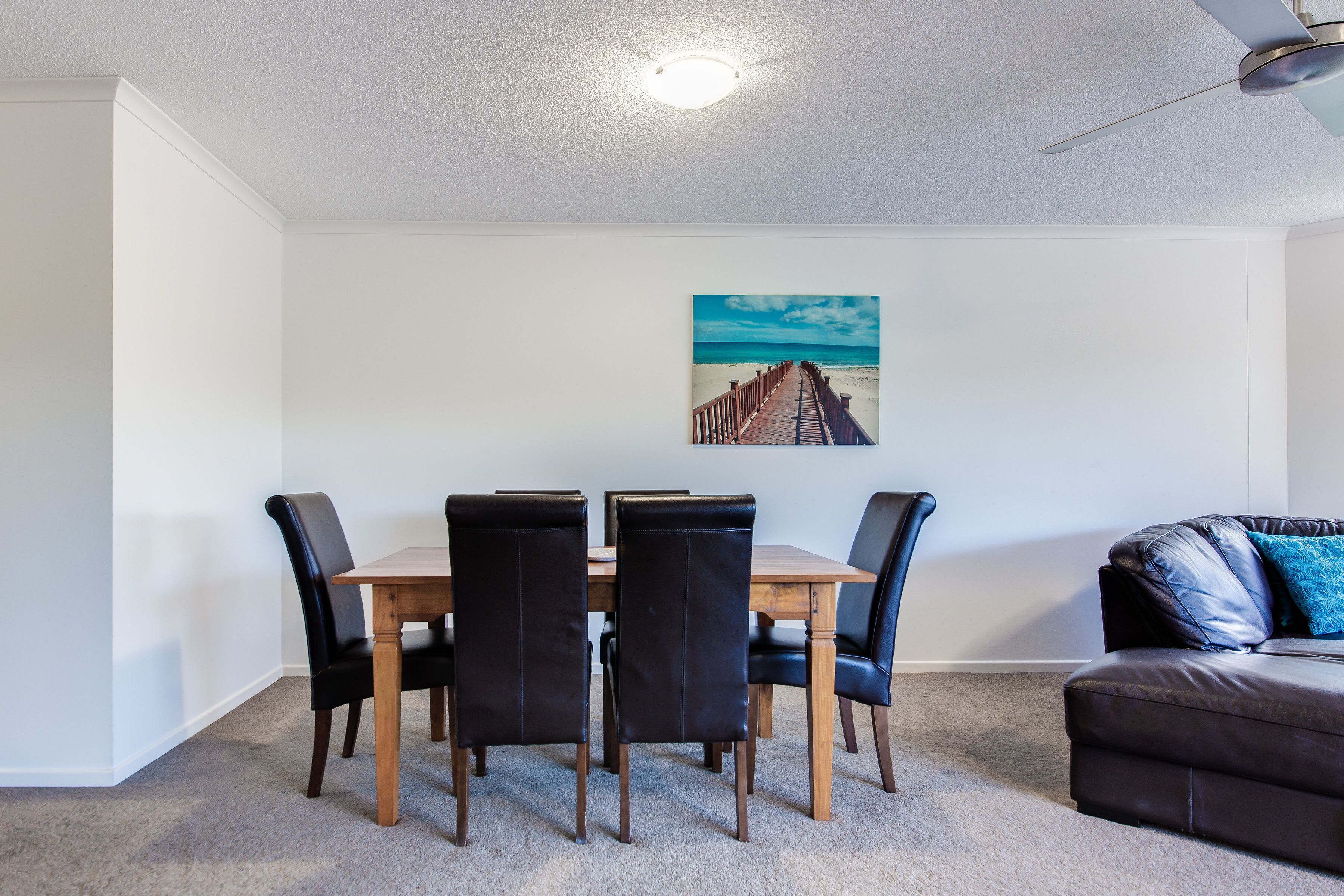 Ein Essbereich mit Holztisch und schwarzen Stühlen, einem Deckenventilator und einem Wandbild mit Strandmotiv.