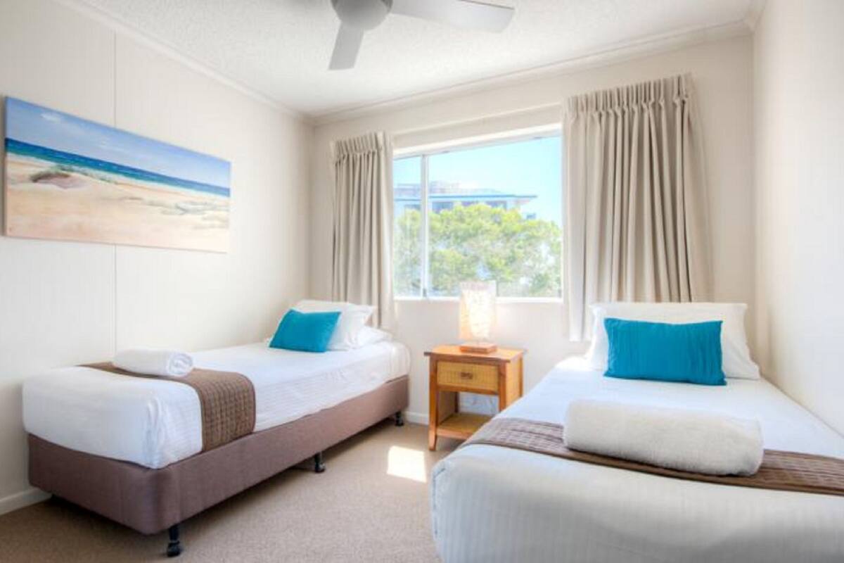 A hotel room with two beds, a painting of a beach, a window with curtains, and a ceiling fan.