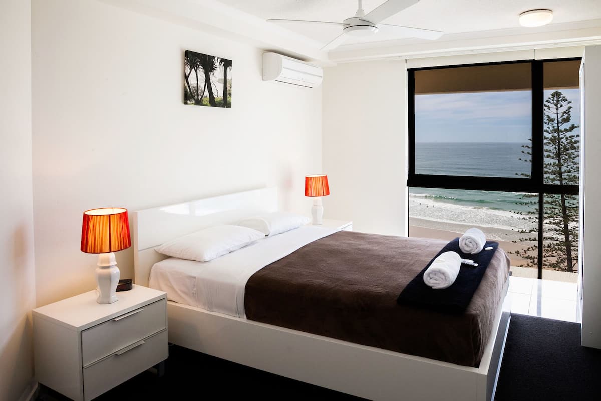 A modern hotel room with a large bed, two bedside lamps, a nightstand, and a view of the beach.
