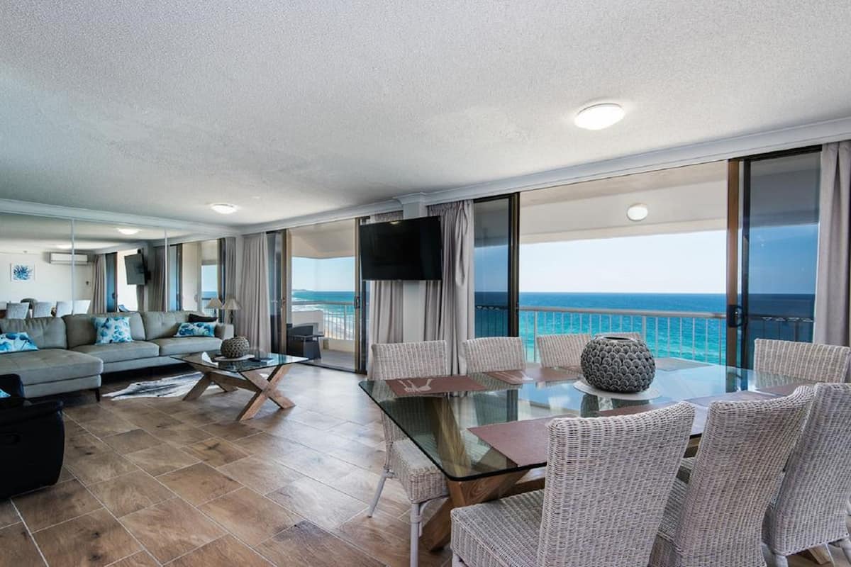 A spacious living area with a glass table and wicker chairs, a sofa with blue cushions, and a large TV.