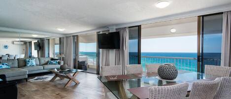 A spacious living area with a glass table and wicker chairs, a sofa with blue cushions, and a large TV.